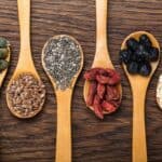 Five wooden spoons on a wooden surface, each holding different seeds and dried fruits: pumpkin seeds, chia seeds, flaxseeds, dried goji berries, dried blueberries, and quinoa.