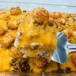 A spatula lifts a portion of a baked casserole featuring tater tots, ground beef, and melted cheddar cheese from a glass dish.