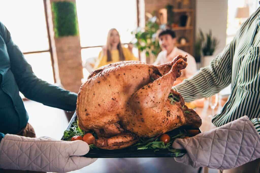 Two people hold a roasted turkey on a tray, garnished with vegetables. In the background, three people sit at a table in a warmly lit room.