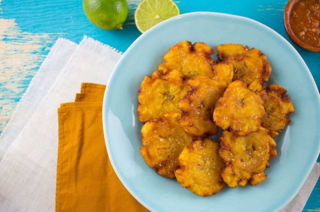 A blue plate with fried plantains, accompanied by a cut lime, a whole lime, and a small bowl of sauce on a wooden surface with yellow and white napkins showcases the culinary delights of the Dominican Republic.