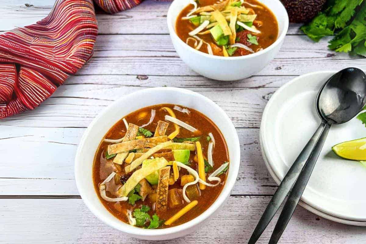 Two bowls of Turkey Tortilla Soup with a plate and spoons beside a wooden table.