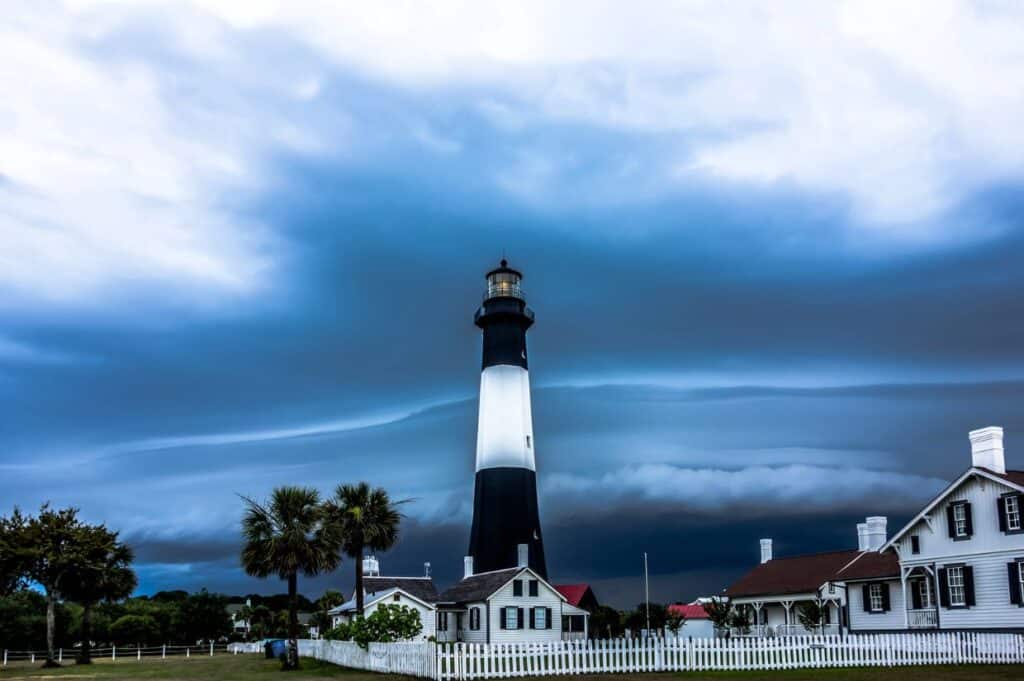 A tall, black-and-white lighthouse stands under a dramatic, cloudy sky, surrounded by several small buildings, a white picket fence, and a few palm trees.