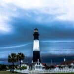 A tall, black-and-white lighthouse stands under a dramatic, cloudy sky, surrounded by several small buildings, a white picket fence, and a few palm trees.