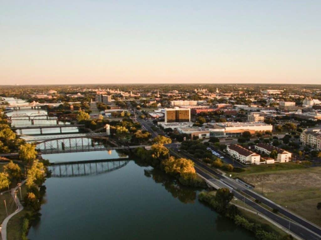 Aerial view of Waco reveals a city with bridges over the meandering river, roads weaving among buildings and lush greenery, all under a clear sky. Discover things to do in Waco while enjoying this picturesque blend of urban life and nature.