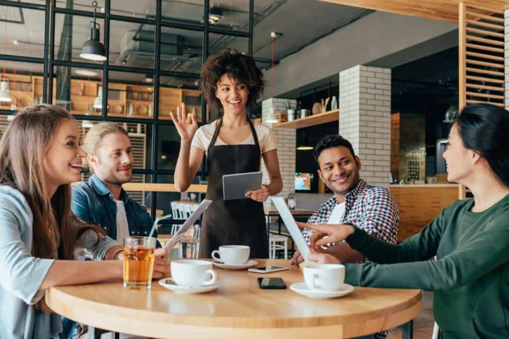 A server greets four people sitting at a table in a cafe. Three have cups of coffee; one has a glass of iced tea. They appear to be enjoying a conversation.