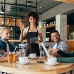 A server greets four people sitting at a table in a cafe. Three have cups of coffee; one has a glass of iced tea. They appear to be enjoying a conversation.