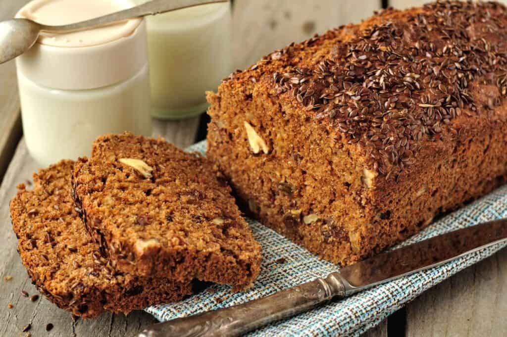 Sliced loaf of brown bread with seeds on top. Two jars with white lids are in the background, and a knife rests nearby on a cloth.