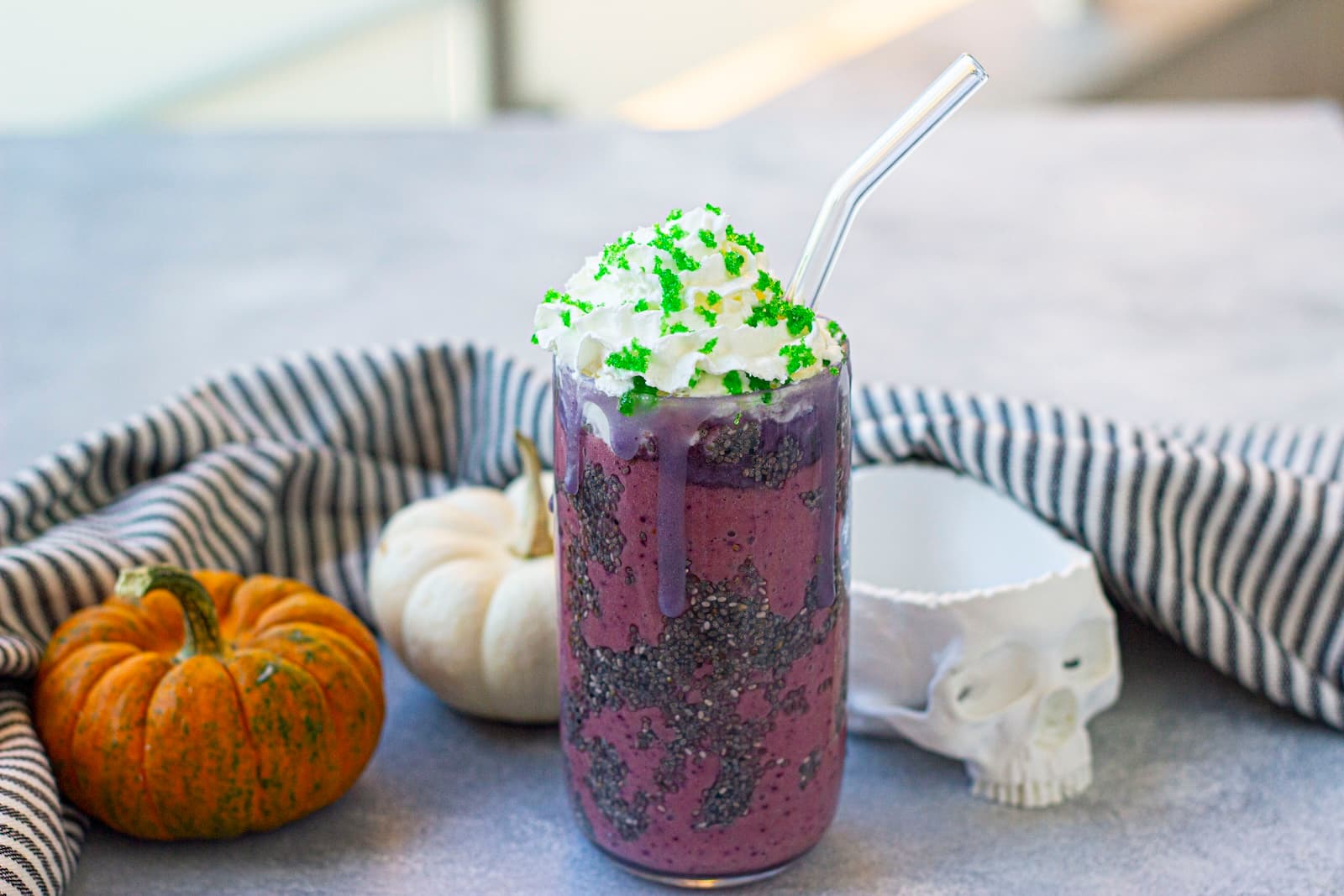 A purple Halloween drink in a glass with whipped cream and green sprinkles, a striped straw, surrounded by two small pumpkins, a striped cloth, and a decorative skull on a gray surface.