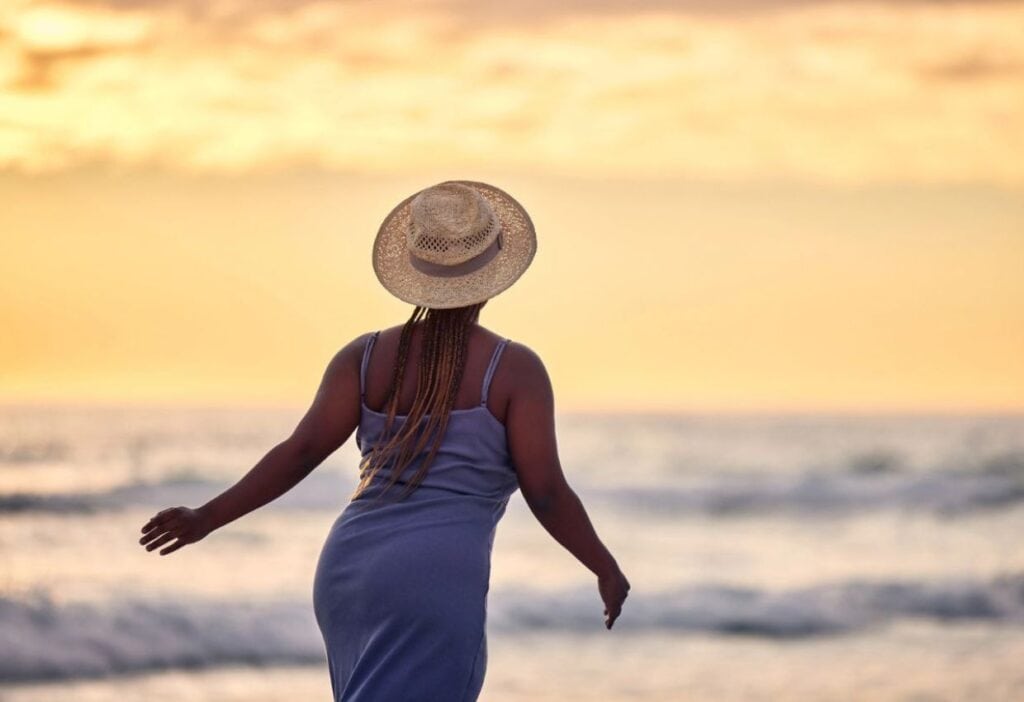 Person in a blue dress and straw hat stands on a beach, facing the ocean, during sunset.