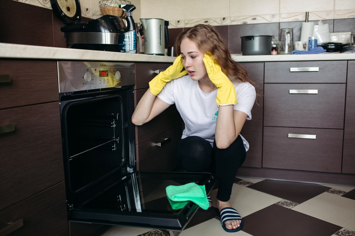 A woman wearing yellow gloves kneels in front of an open oven in a modern kitchen, appearing frustrated. A green cloth lies on the floor, and various kitchen items are on the counter.