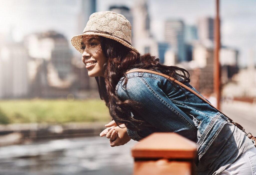 A woman in a denim jacket and straw hat is leaning on a bridge railing, smiling, with a cityscape in the background.