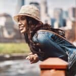 A woman in a denim jacket and straw hat is leaning on a bridge railing, smiling, with a cityscape in the background.