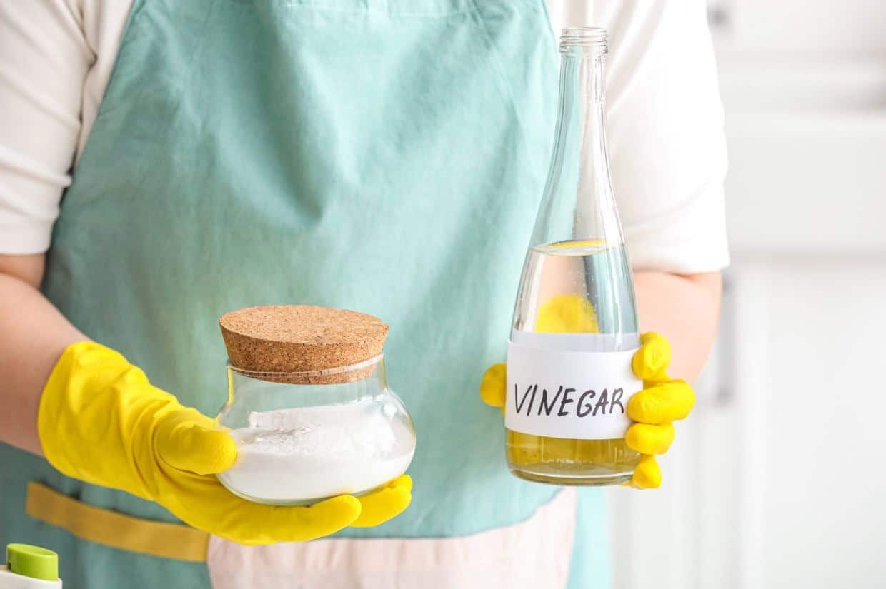 Person in apron and yellow gloves holding a jar of baking soda and a bottle labeled vinegar.