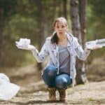 A woman kneels in the forest holding a plastic bottle in one hand and crumpled paper in the other, with a confused expression. A white garbage bag is on the ground beside her.