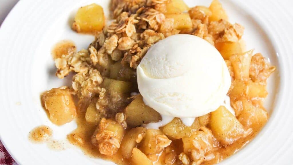 A plate of apple crisp with chunks of apple, topped with a scoop of vanilla ice cream and an oat crumble topping.