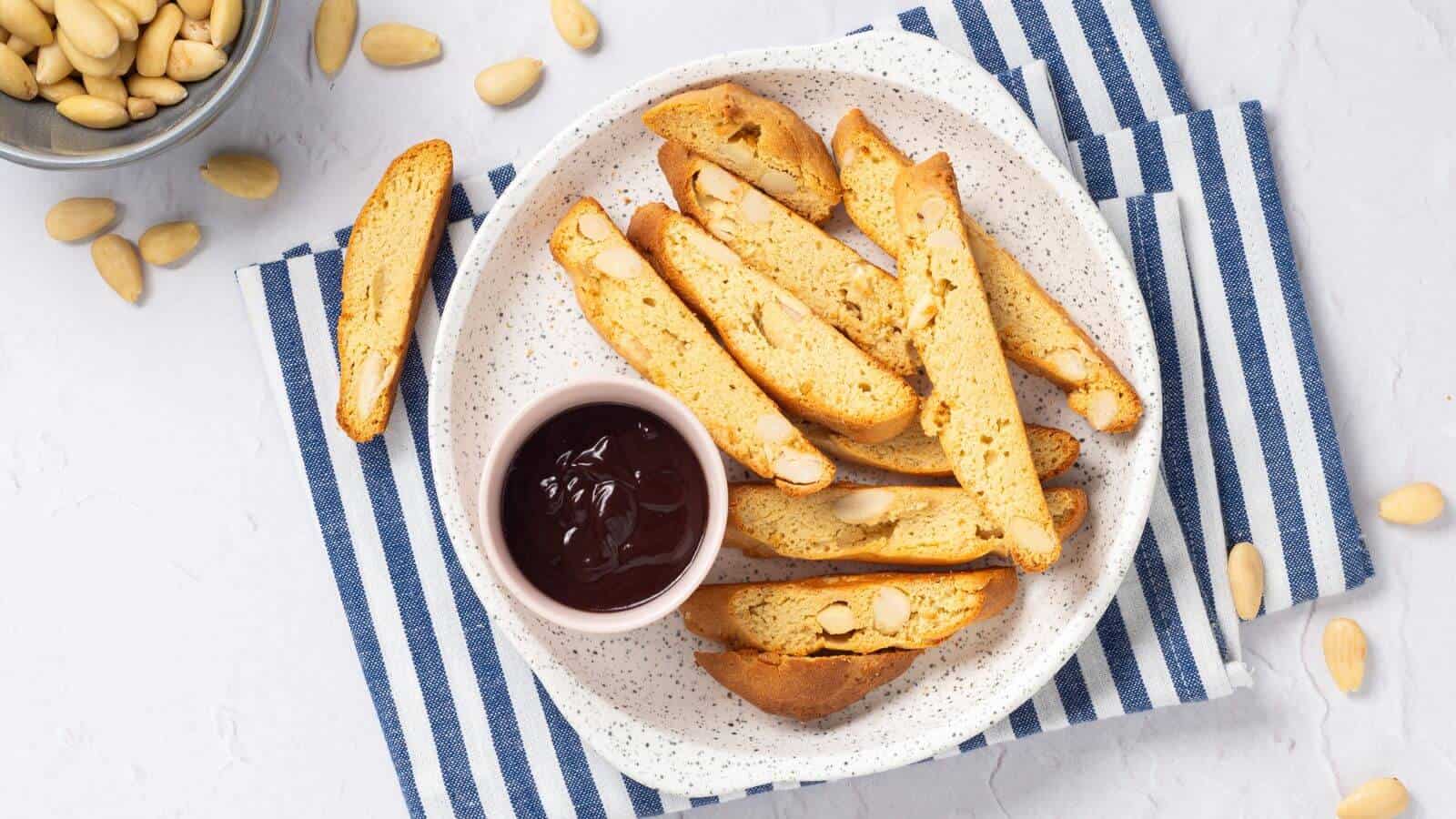 A plate of almond biscotti with a small bowl of chocolate sauce on a blue and white striped cloth. Almonds are scattered around.