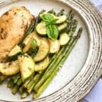 Grilled chicken breast with zucchini and asparagus, garnished with basil, served on a speckled ceramic plate.