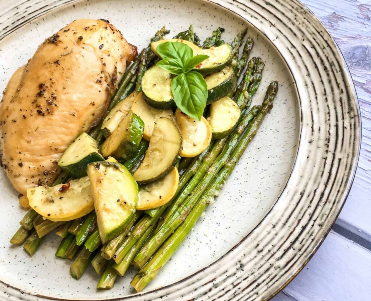 Grilled chicken breast with zucchini and asparagus, garnished with basil, served on a speckled ceramic plate.