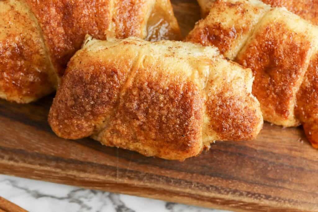 Close-up of golden-brown baked pastry rolls on a wooden board, with a visible flaky texture and sprinkled with cinnamon sugar.