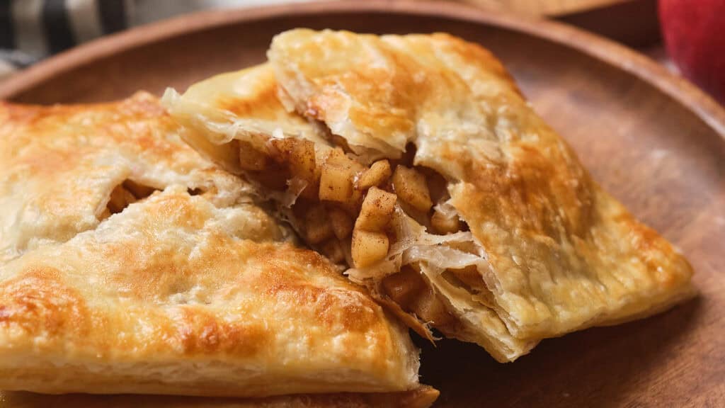 A flaky pastry filled with spiced apple chunks is served on a wooden plate.