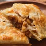 A flaky pastry filled with spiced apple chunks is served on a wooden plate.