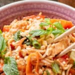 A bowl of stir-fried noodles with vegetables, chicken, chopped peanuts, and fresh mint, with chopsticks resting on the rim.