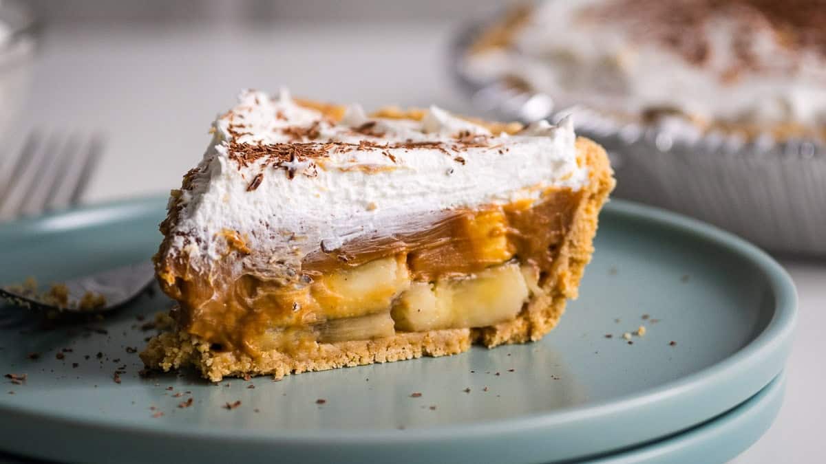 A slice of banoffee pie on a light blue plate, featuring layers of banana, caramel, and whipped cream, with a sprinkle of chocolate shavings on top.