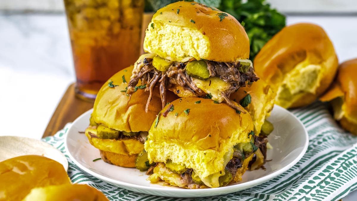 Three pulled pork sliders with pickles and cheese are stacked on a white plate.