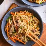 A bowl of stir-fried noodles with vegetables and meat, being picked up with chopsticks. Mushrooms and sesame seeds are visible nearby.