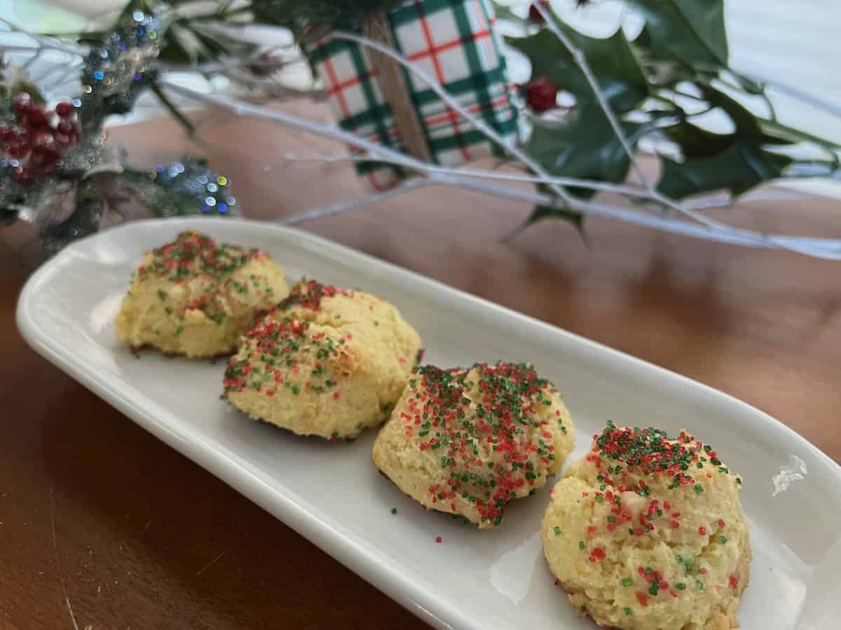 Four scones topped with red and green sprinkles on an oval plate. They sit on a wooden table with holiday decorations in the background.