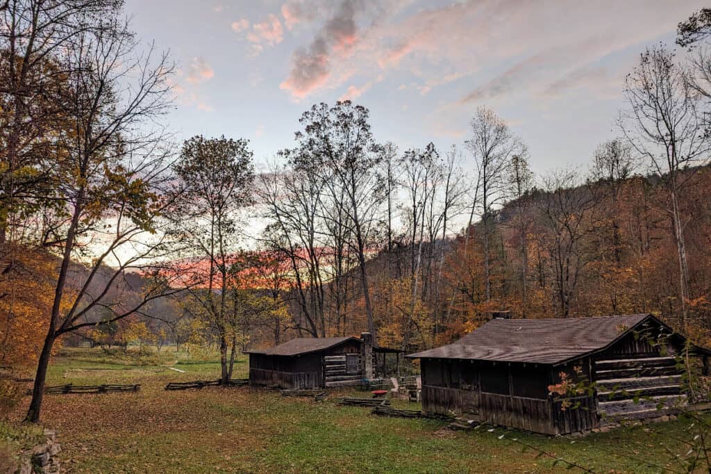 Two rustic log cabins in a forest clearing at sunset with trees showing fall foliage and mountains in the background.