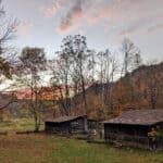 Two rustic log cabins in a forest clearing at sunset with trees showing fall foliage and mountains in the background.