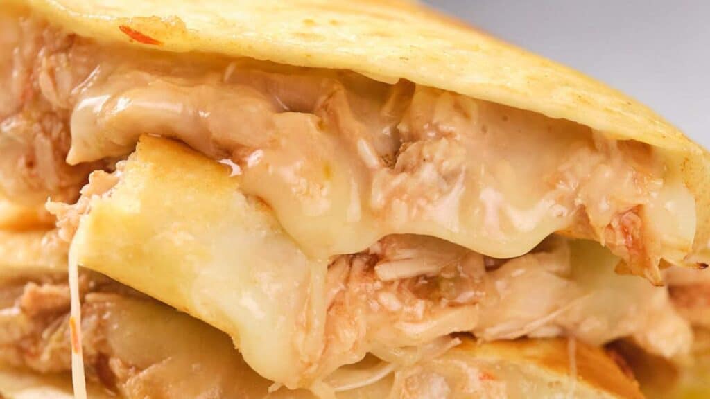 Close-up of a cheesy quesadilla with pulled chicken and melted cheese oozing between crispy, golden-brown flour tortillas.