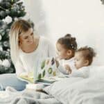 A woman and two children read a book together in a cozy room with a decorated Christmas tree.