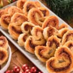 A rectangular white platter and a small bowl filled with heart-shaped palmiers, surrounded by holiday decorations, including red and gold baubles and green foliage.