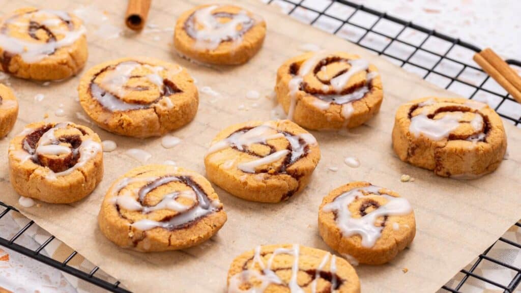 Small cinnamon rolls with icing are placed on a sheet of parchment paper on a cooling rack. Two cinnamon sticks are visible beside them.