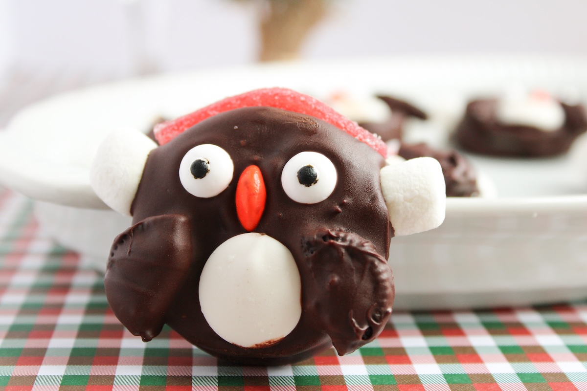 A chocolate-covered treat shaped like a penguin, with candy eyes, a red beak, and white marshmallow wings, on a checkered tablecloth.