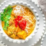 A bowl of noodle soup with chicken, garnished with cilantro, sliced red chili, and a dollop of red sauce. Another smaller bowl for garnish is on the side.