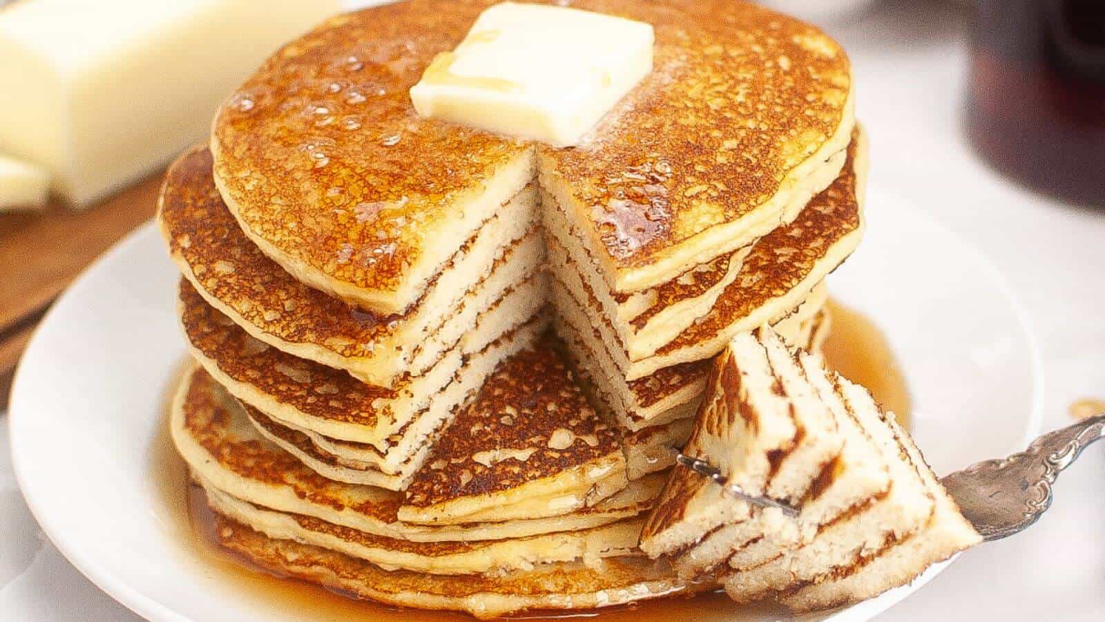 A stack of pancakes with a pat of butter on top, drizzled with syrup. A wedge has been cut out and is resting on a fork beside the stack.