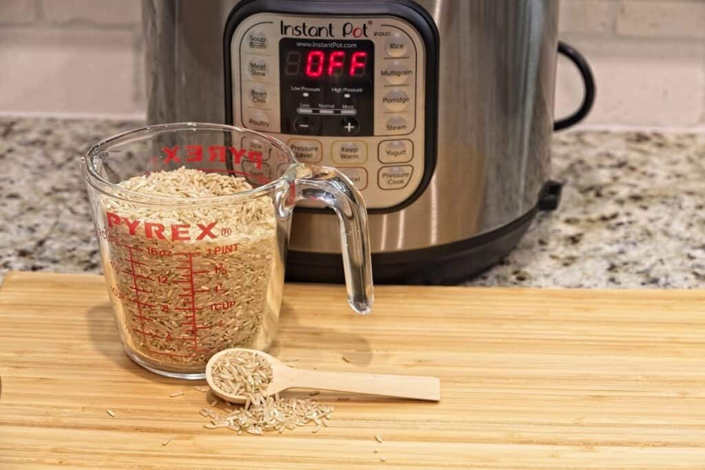 A measuring cup filled with uncooked brown rice sits on a wooden board in front of an Instant Pot displaying "OFF.