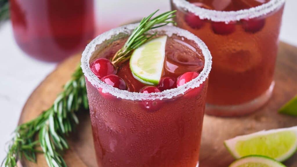 Two glasses filled with a red beverage, garnished with cranberries, lime slices, and rosemary sprigs, sit on a wooden surface. The rims of the glasses are coated in sugar. Lime slices are nearby.