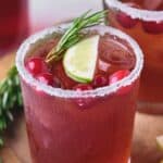 Two glasses filled with a red beverage, garnished with cranberries, lime slices, and rosemary sprigs, sit on a wooden surface. The rims of the glasses are coated in sugar. Lime slices are nearby.