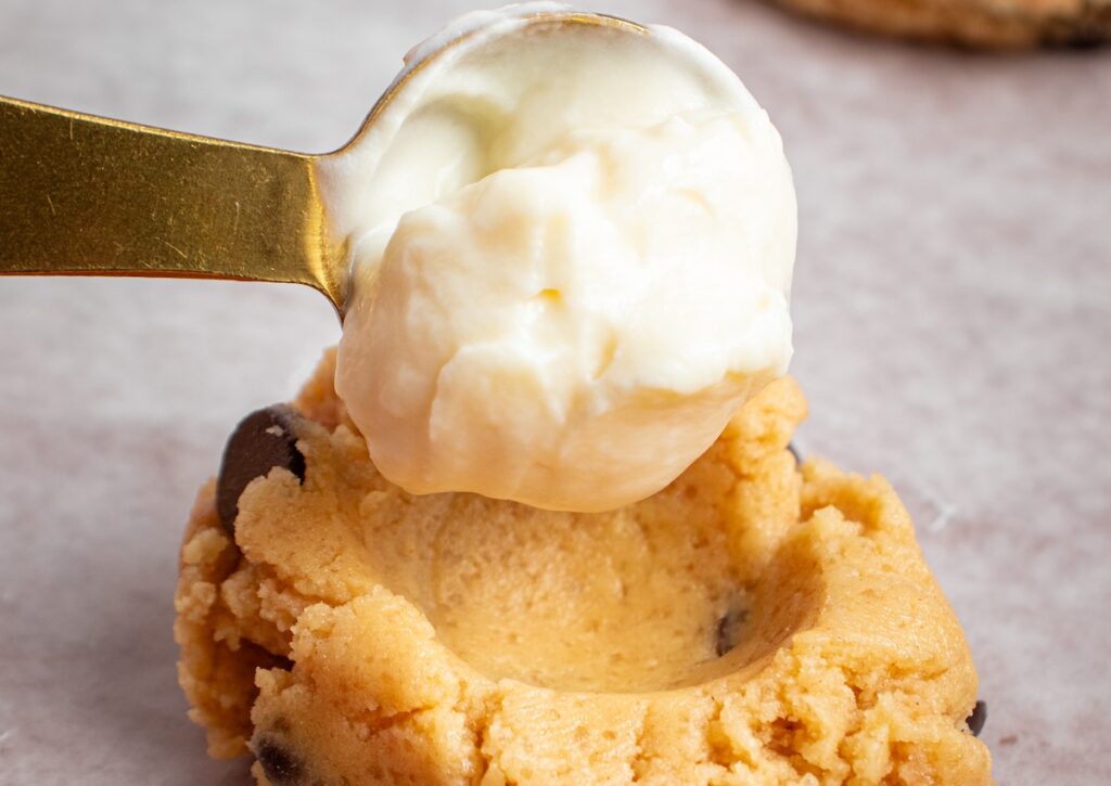 A scoop of cream cheese is being placed into a cookie dough indentation.