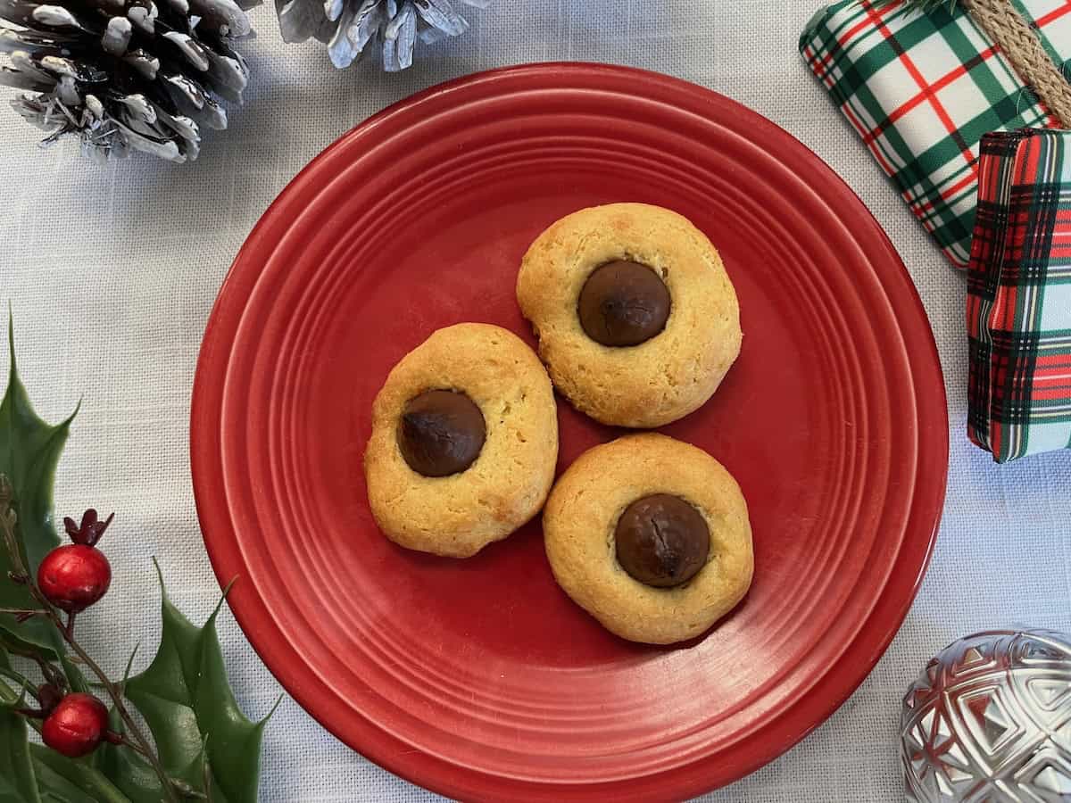 Three cookies with chocolate centers on a red plate, surrounded by festive decorations including pinecones and wrapped gifts.