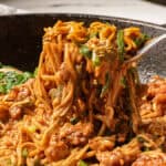 A fork lifts saucy noodles with ground meat and spinach from a skillet.