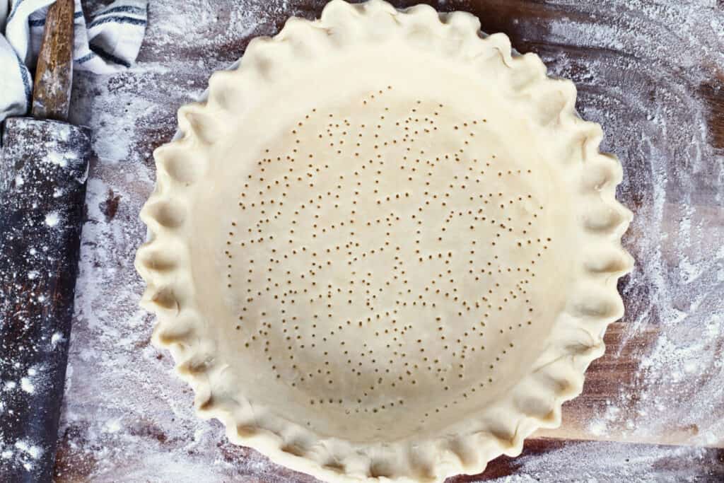 Unbaked pie crust in a pan with crimped edges, pricked with a fork. Rolling pin and flour-dusted surface visible in the background.