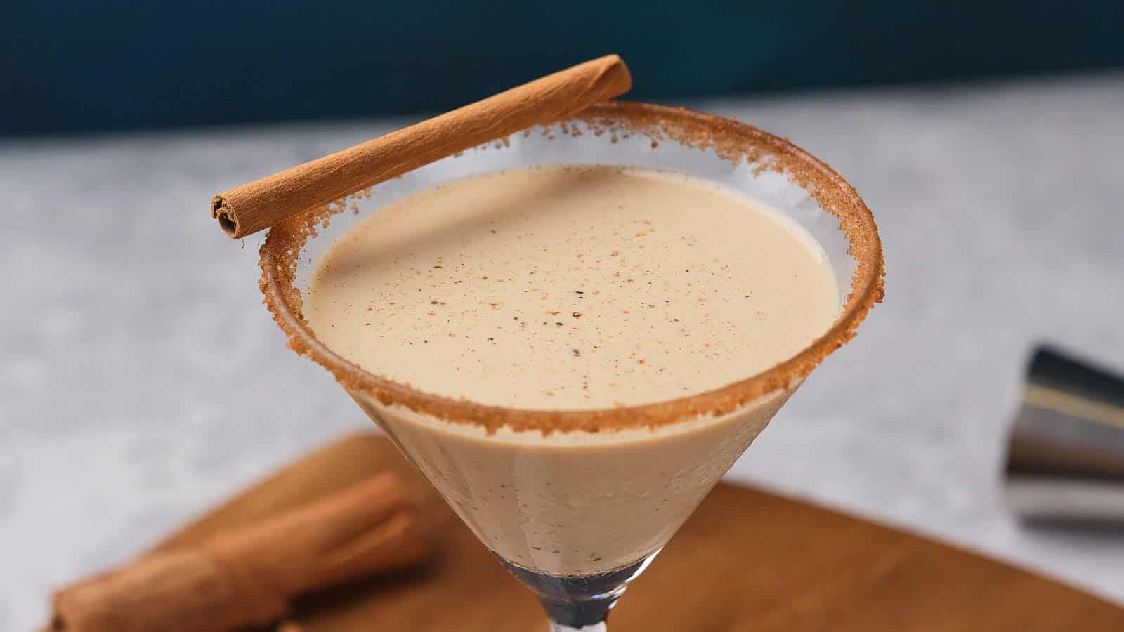 Eggnog martini in a glass, garnished with a sugar rim and cinnamon stick, placed on a wooden board.