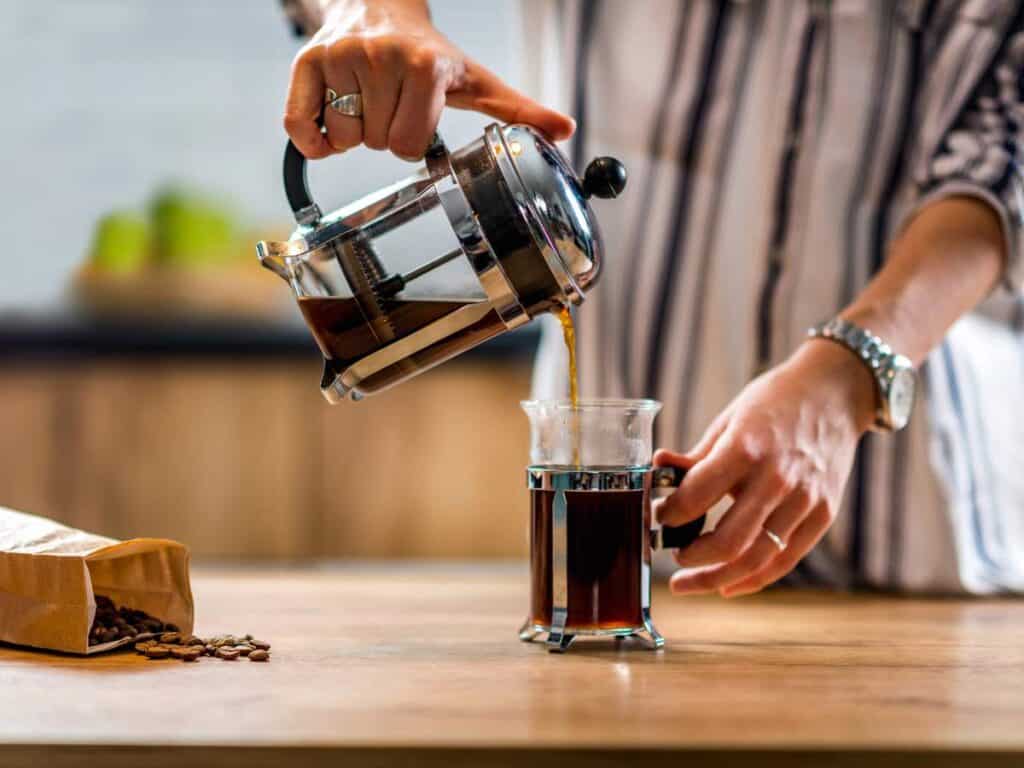 A person pours coffee from a French press into a glass cup on a wooden table, creating the perfect cozy touch for a fall breakfast.