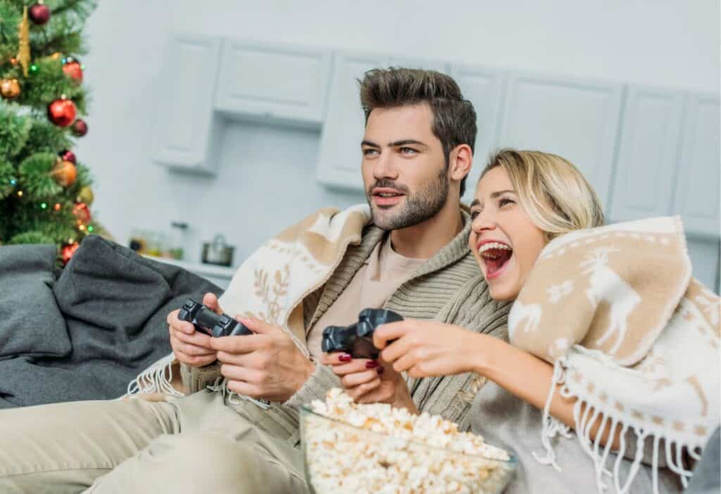 A man and woman sit on a sofa covered with blankets, playing video games and smiling. A bowl of popcorn is in front of them, with a decorated Christmas tree in the background.