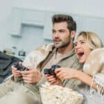 A man and woman sit on a sofa covered with blankets, playing video games and smiling. A bowl of popcorn is in front of them, with a decorated Christmas tree in the background.